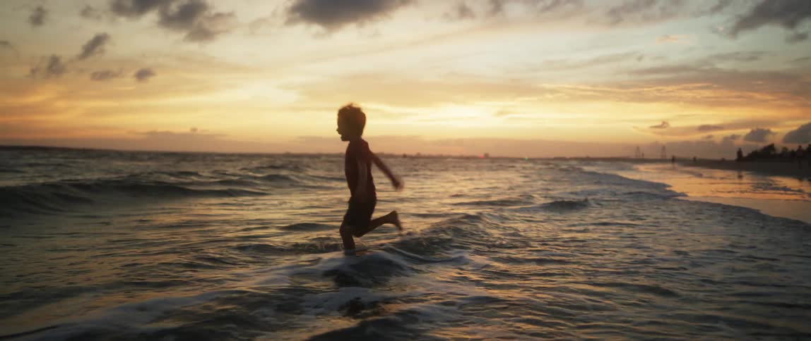 A young boy on the beach jumps over waves rolling in and then turns and runs to adults standing on the beach
