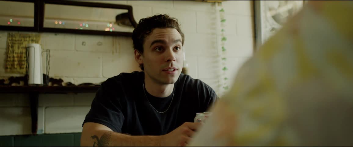A man is sitting in a restaurant holding a beer can and talking to another person