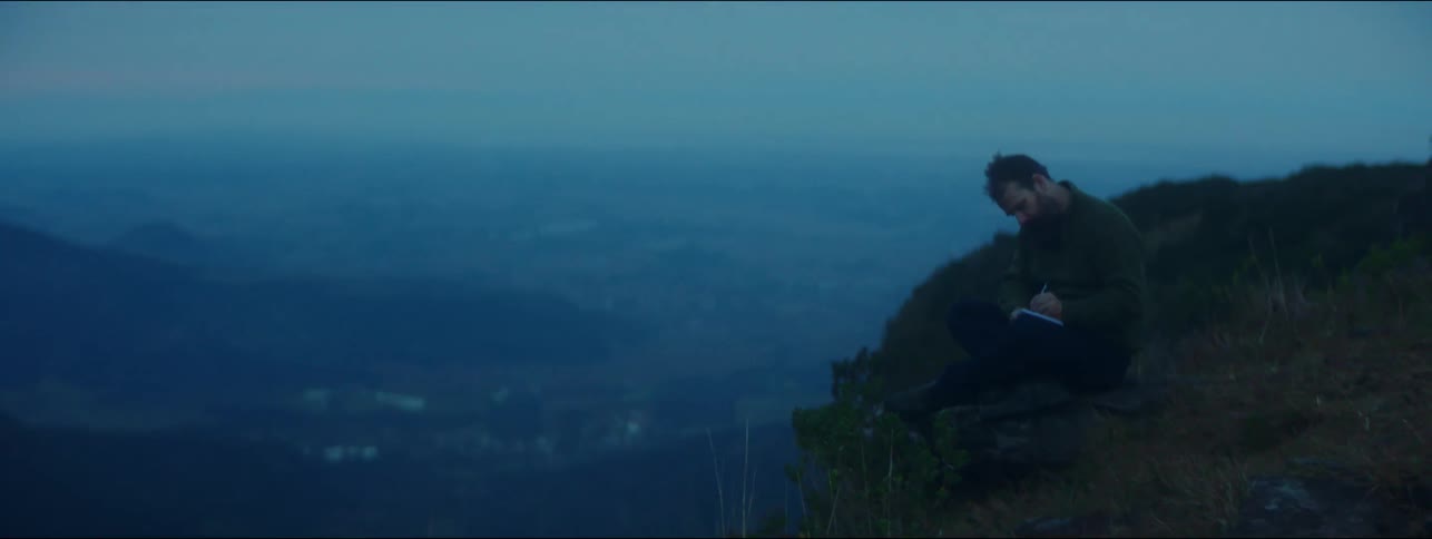 A man writes in a notebook while sitting on the edge of a cliff