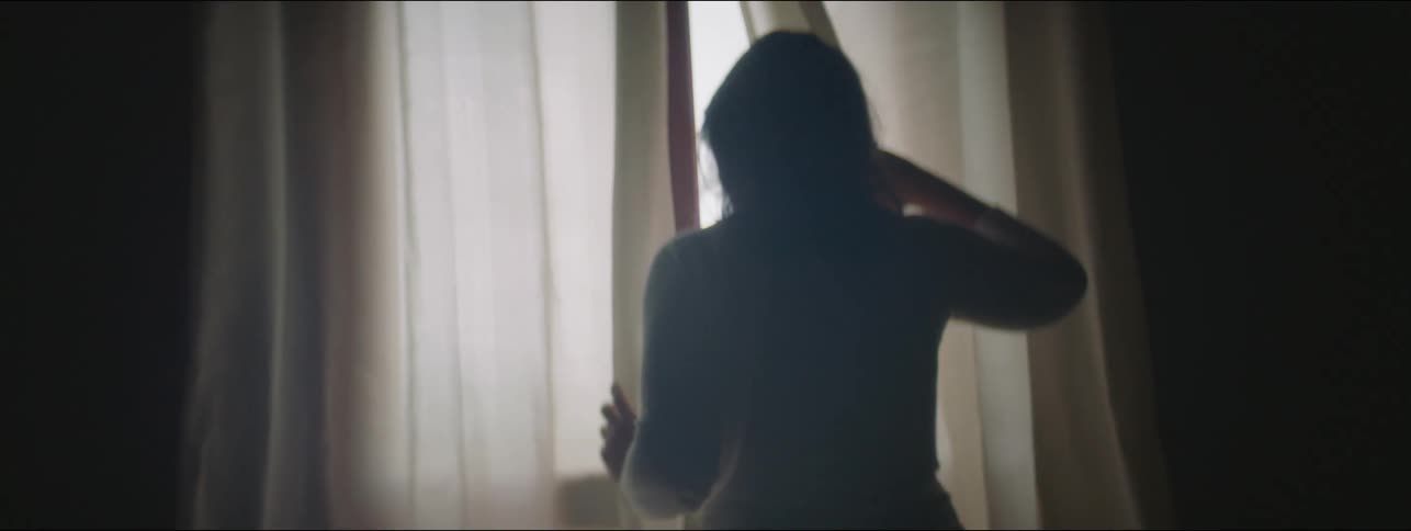 A woman at home walks up to the window and pulls the curtain back and peeks outside