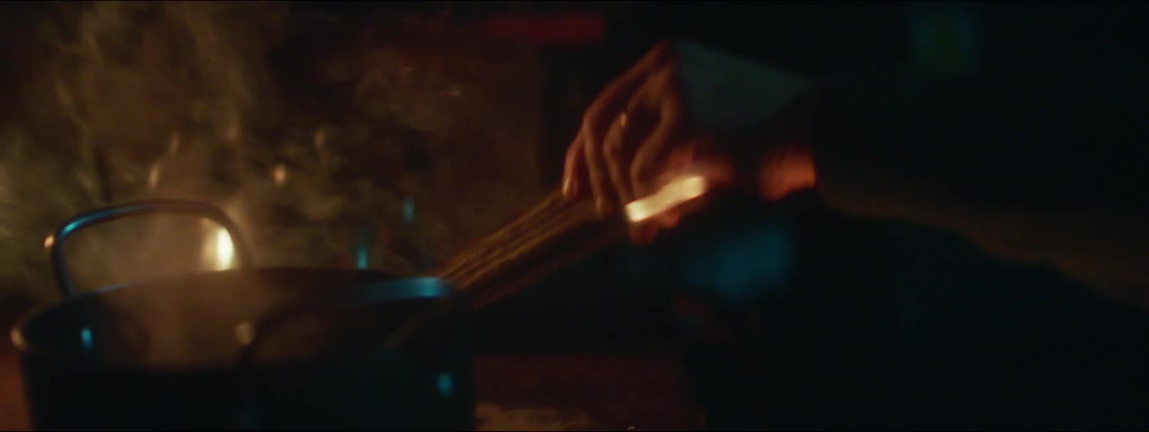 A person uses a piece of wood to stoke the flames over an old kitchen inside a dimly lit cabin