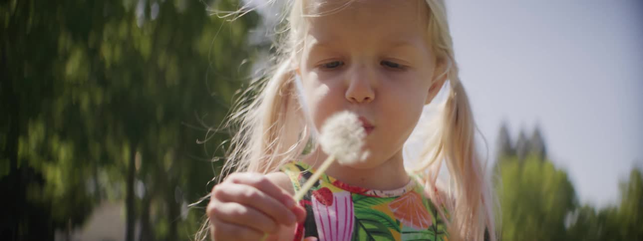 Blonde girl stands outside blowing dandelion seeds