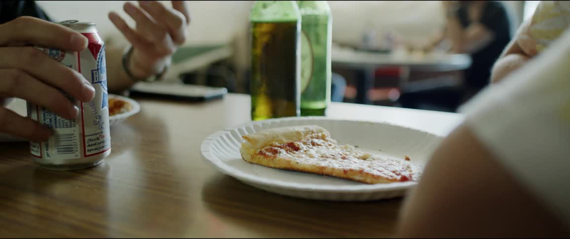 A slice of pizza lies on a paper plate on a table between two people chatting in a restaurant