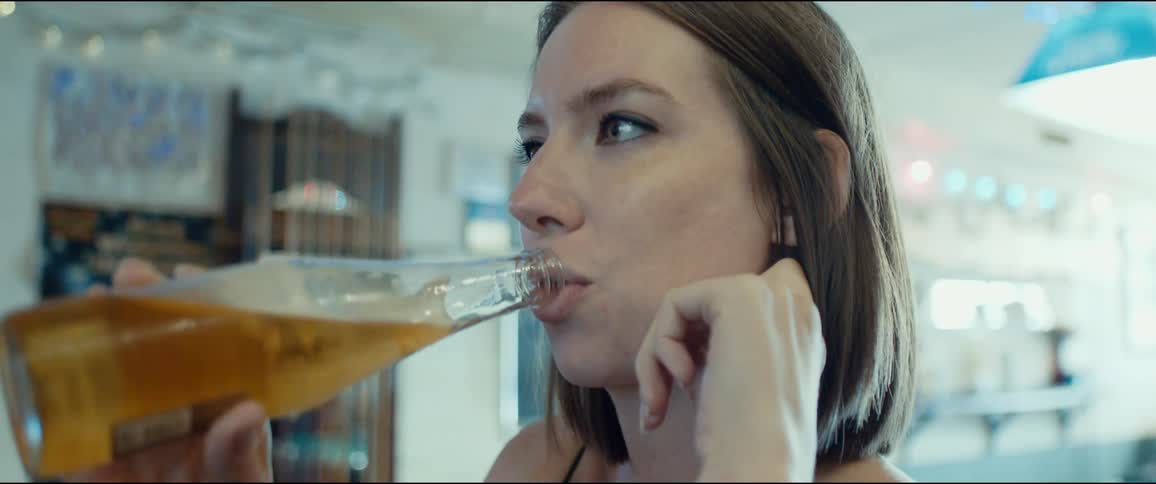 A woman drinks beer from a bottle while talking with a companion in a restaurant during the day