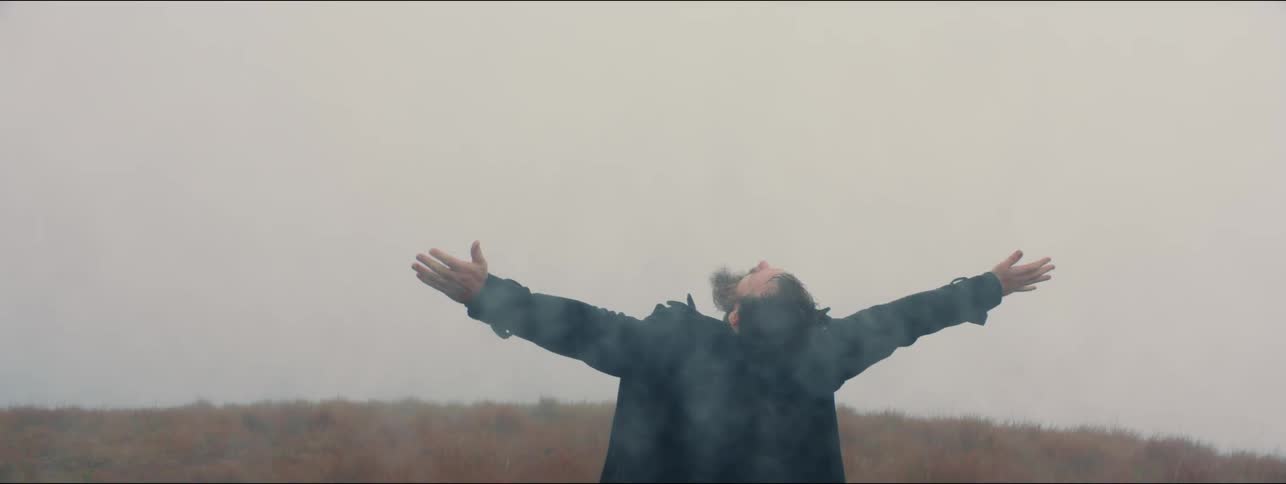 Man wearing jacket outdoors near hill reaches his arms out and bends backward