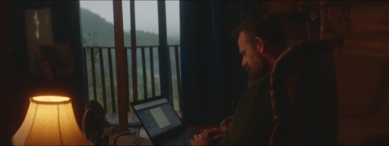 A man is sitting in a chair in a living room typing on a laptop and smiling