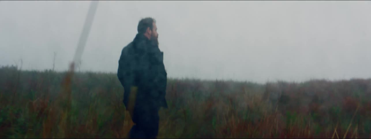Man wearing coat walks through overgrown field in rainy day