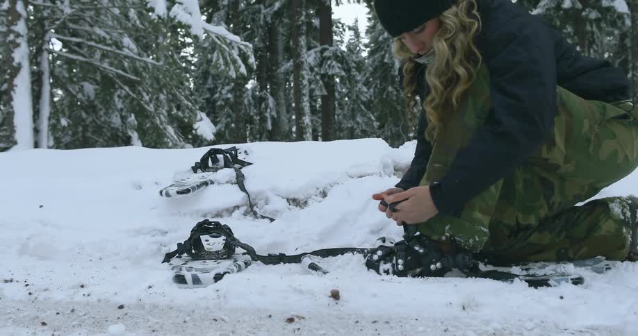 A woman kneels on the ground putting on her snowshoes