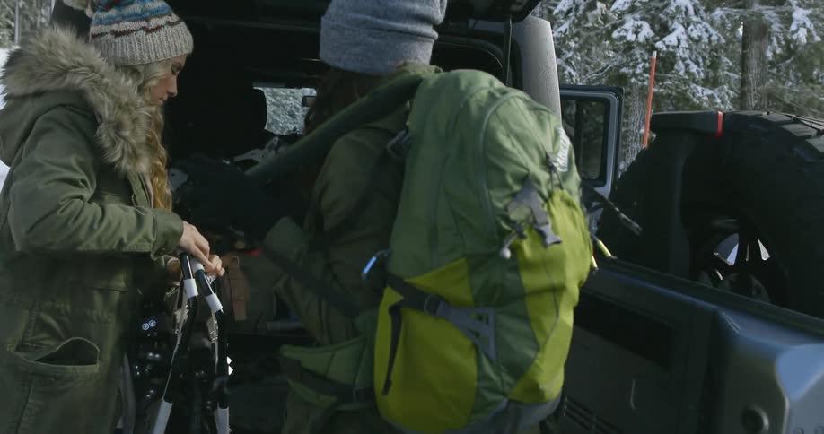 Two woman put on backpacks and grab gear out of a vehicle