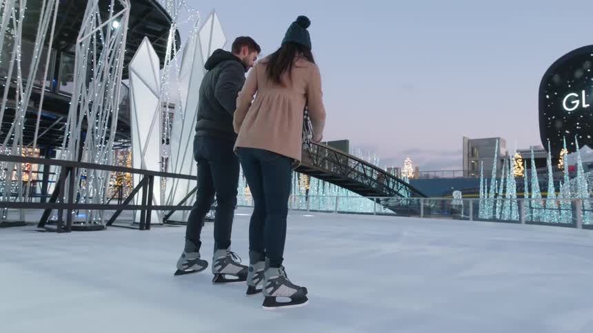 Couple ice skate on outside ice rink decorated with christmas lights