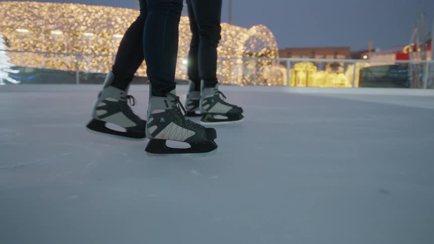 Two people ice skate side by side on an outdoor rink surrounded by tree lights and decorations