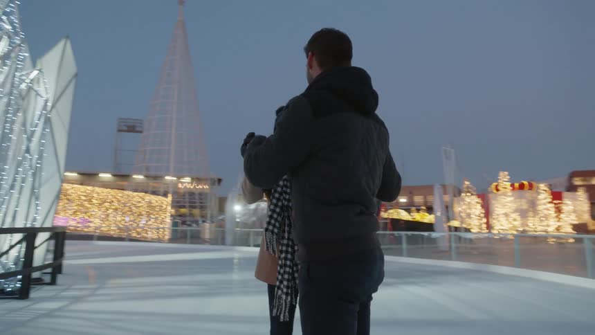 A young couple is holding hands while on ice skates on an outdoor skating rink illuminated by holiday lights