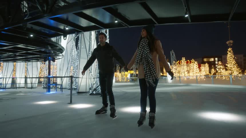 A man and woman hold hands while ice skating atop an outdoor ice rink in a park with many christmas lights and decorations