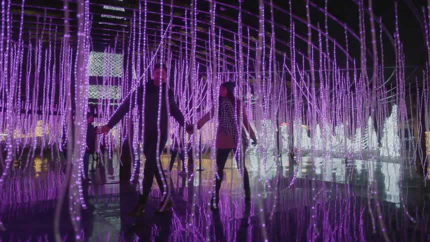 A man leading a woman through hanging purple christmas lights outdoors at night