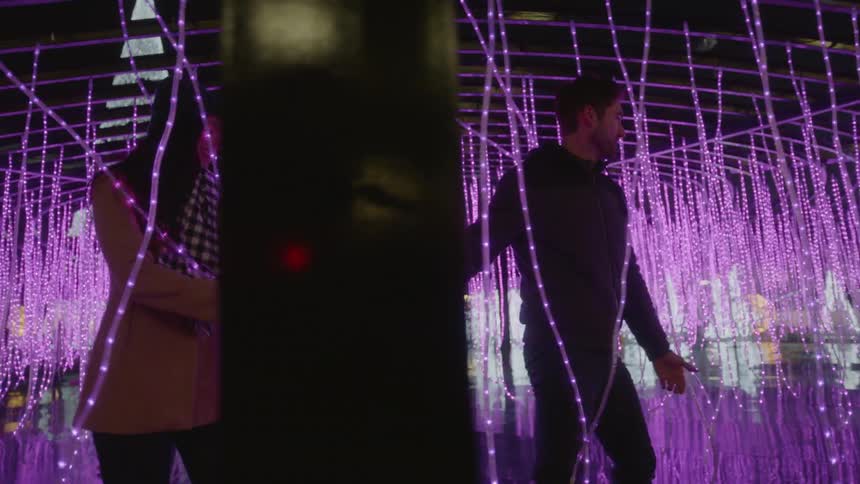 A man and woman hold hands and walk through a light display