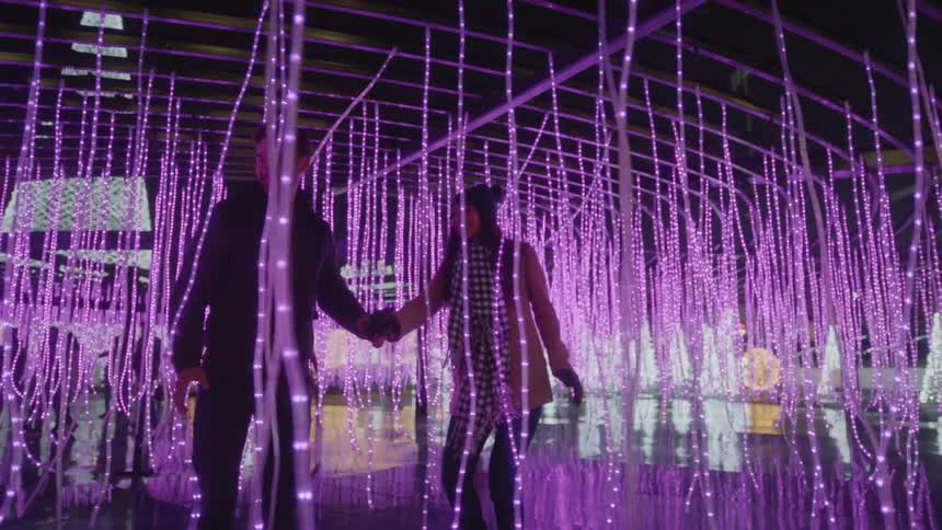 A man leading a woman through an outdoor structure filled with hanging strings of purple light