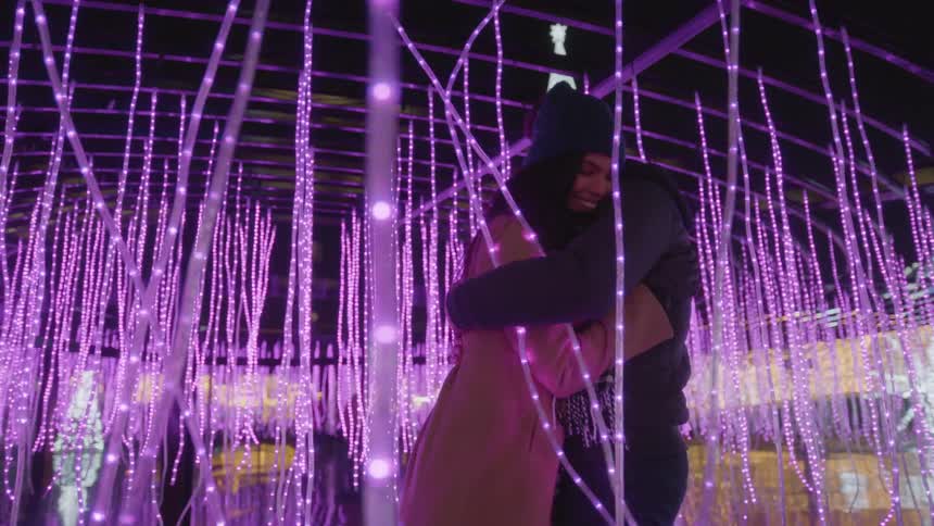 A man and woman hug in an area with several pink hanging string lights
