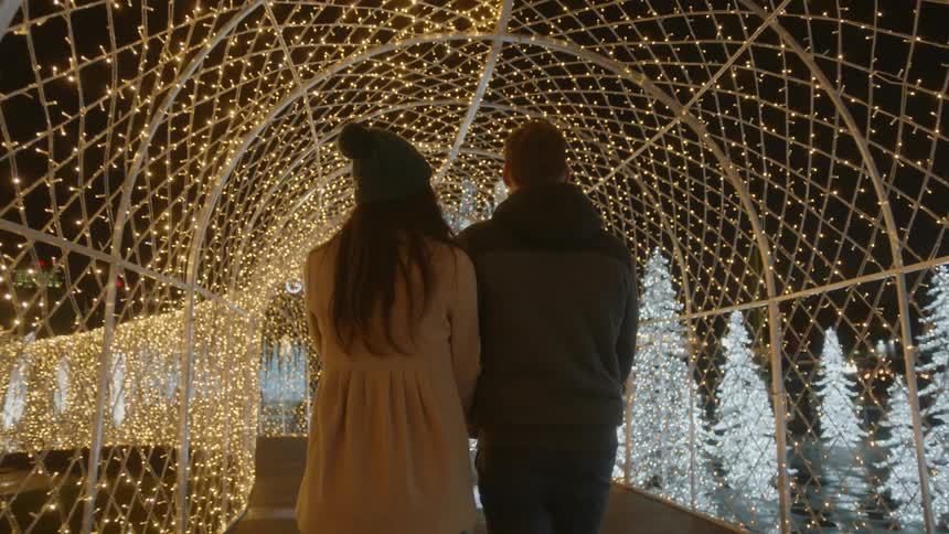 A man and woman walk on a pedestrian bridge that is covered and lit up with yellow lights