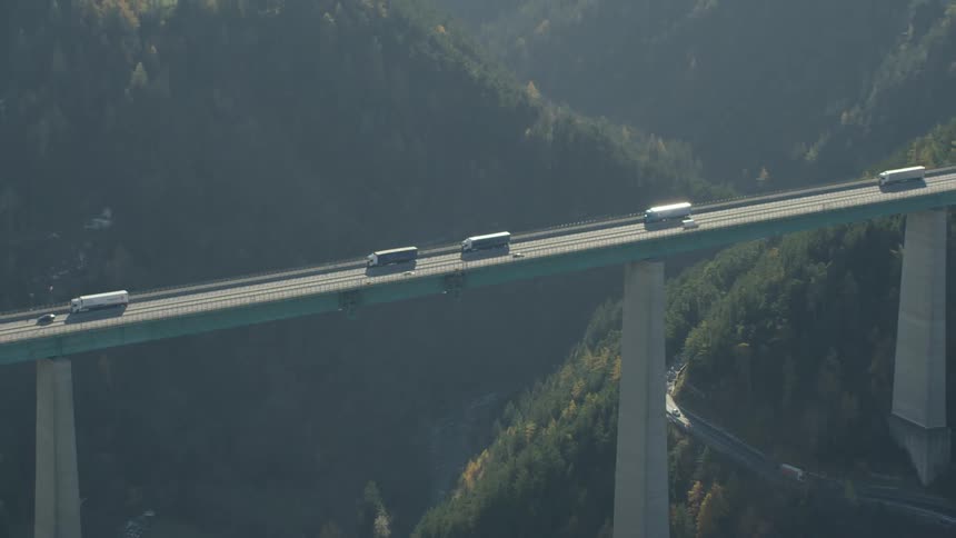 Several semi trucks move along long bridge between mountains in one direction