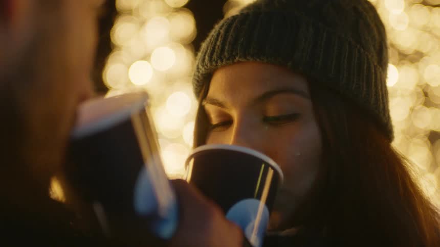 Couple stand outside near christmas lights at night drinking from steaming mugs and smiling