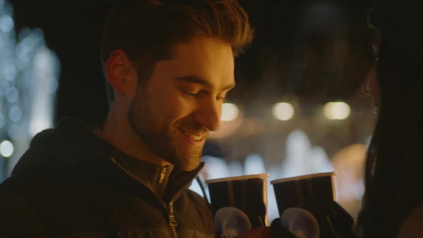A man takes a drink of his coffee while talking with someone else on a cold evening