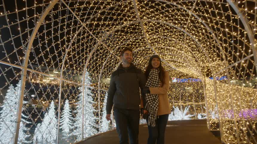 Couple walks through mesh covered tunnel strewn with christmas lights at night