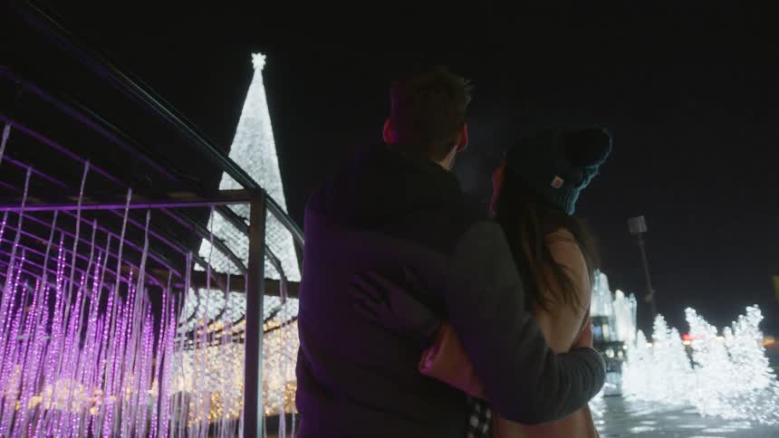 A couple hug in an area illuminated by white and purple holiday lights