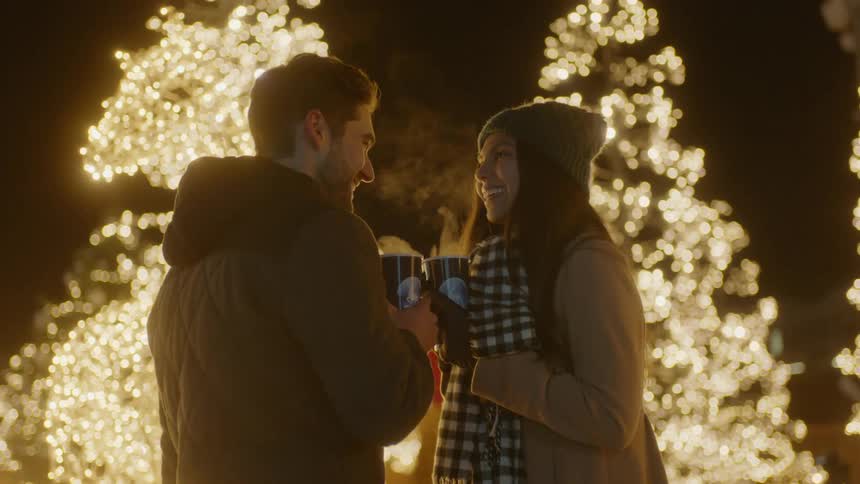 A man and woman standing outdoors in front of a row of trees covered in christmas lights