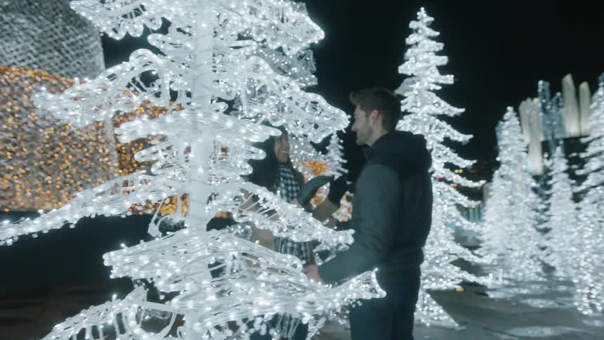 A man runs around a lighted christmas tree display to grab a woman on the other side