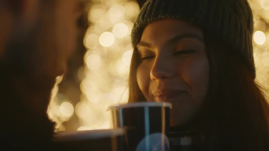 Woman wearing wooly hat drinks coffee and looks at man drinking coffee at night