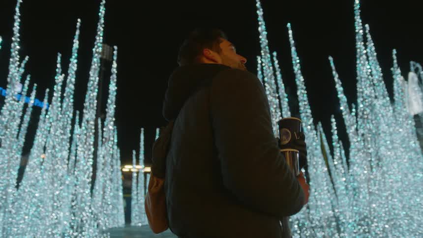 A man hugs his female companion as then stand outside in the middle of a brightly lit plaza decorated to resemble a winter wonderland