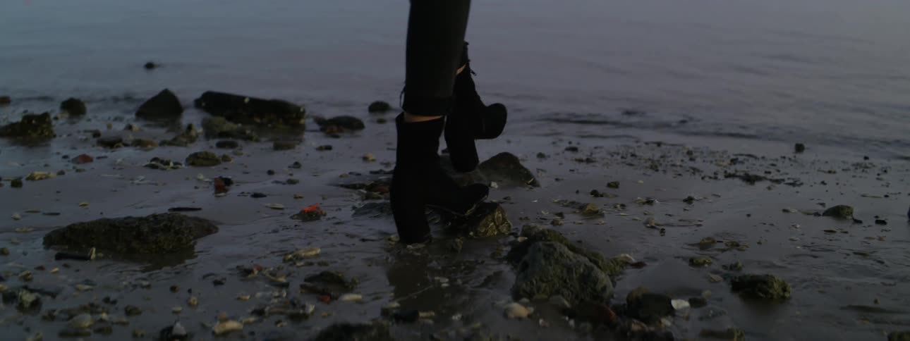 A person wearing high heeled boots walks at the edge of the water