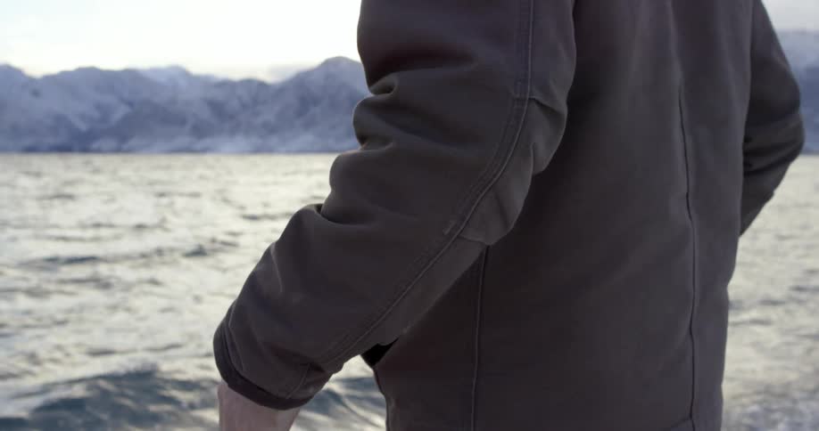 Man with a grey coat puts his hands in his pocket and watches the sea and snow capped mountains on a cold winter day