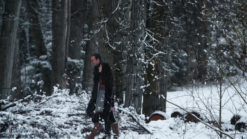 A man and women walk near a forest as it snows outside