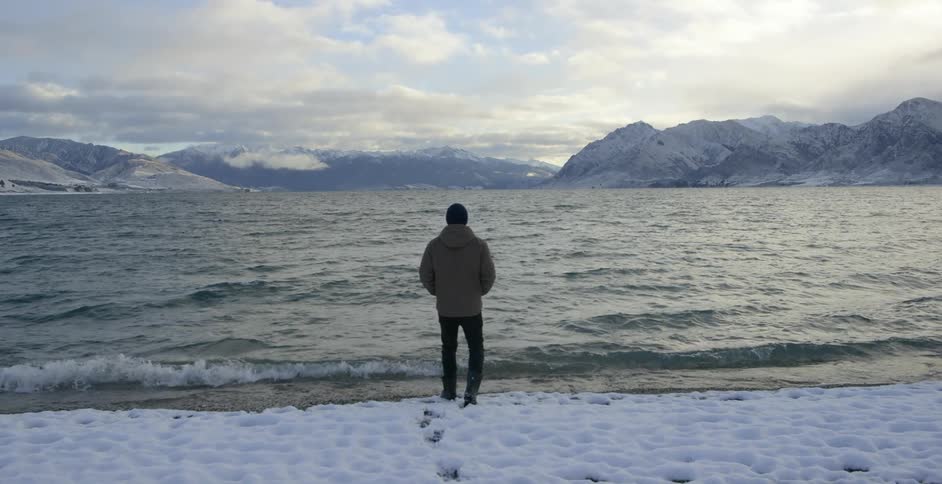 A man walks through the snow towards a mountain lake