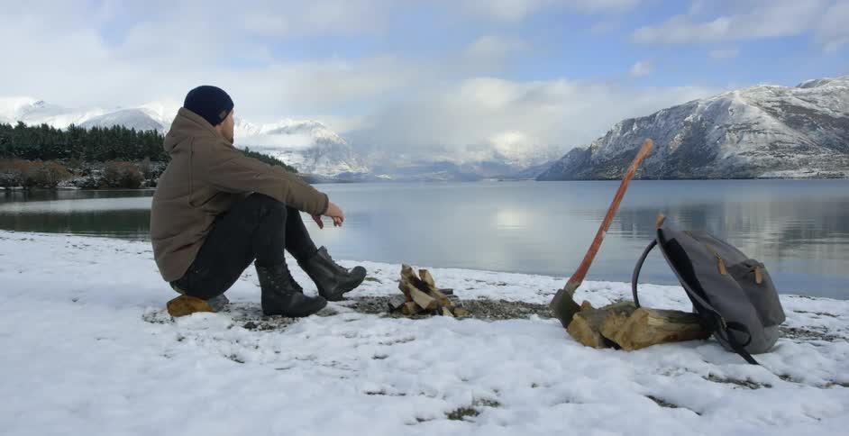 Man sitting in snow in front of a small fire in front of a lake
