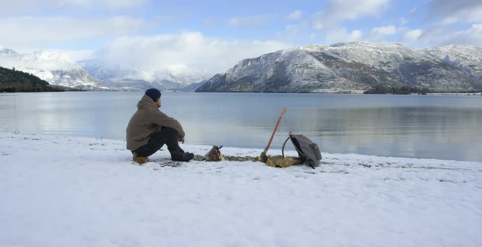 Man sitting on snowy ground in front of a small fire before a lake