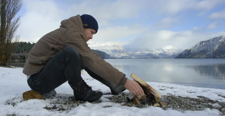 Man sitting on a snowy ground in front of a small fire by a lake