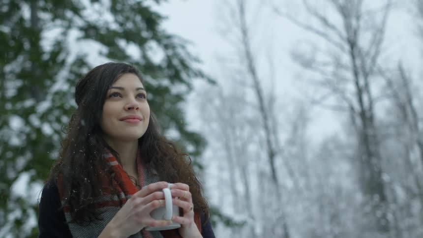 Woman standing outside holding mug smiling as snow falls