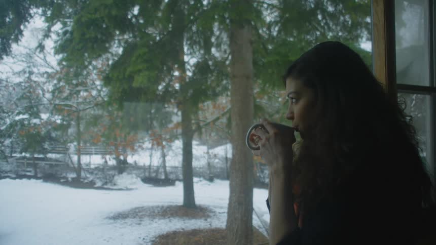 A woman is drinking from a mug as she looks through a window