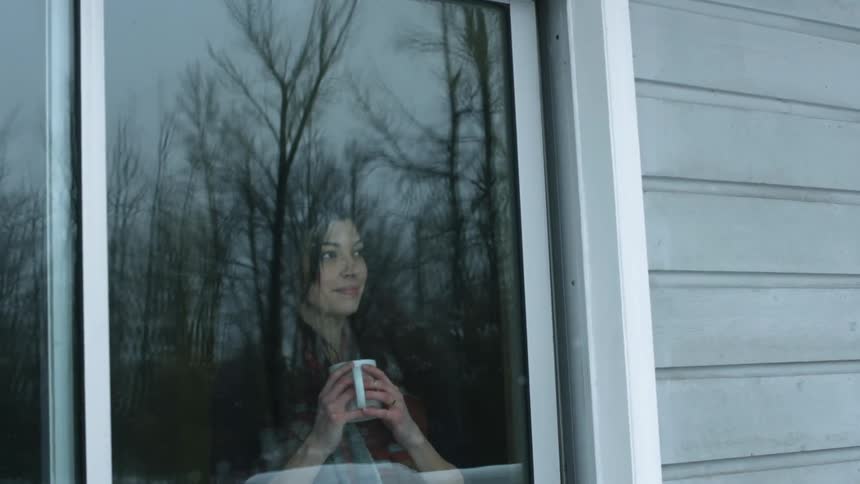 A woman is holding a mug near a window looking out at the snow