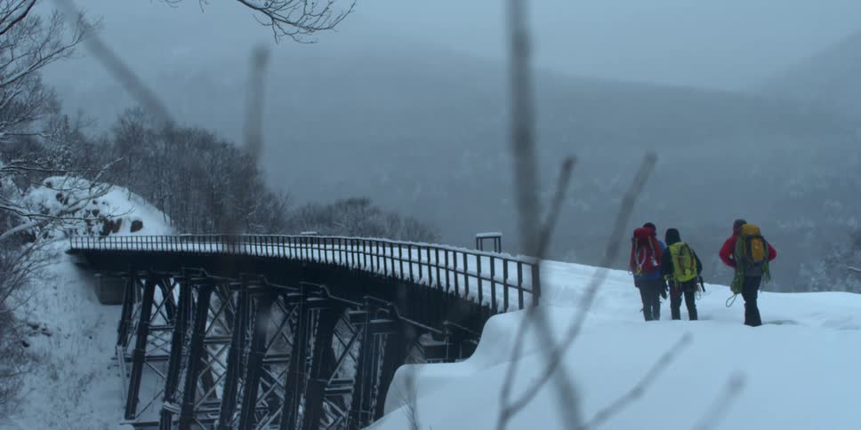 Three people are hiking in the snow across a bridge