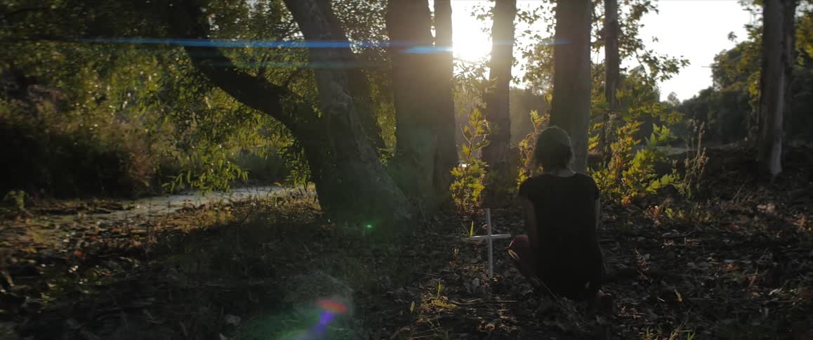 A woman kneels next to a cross in the woods