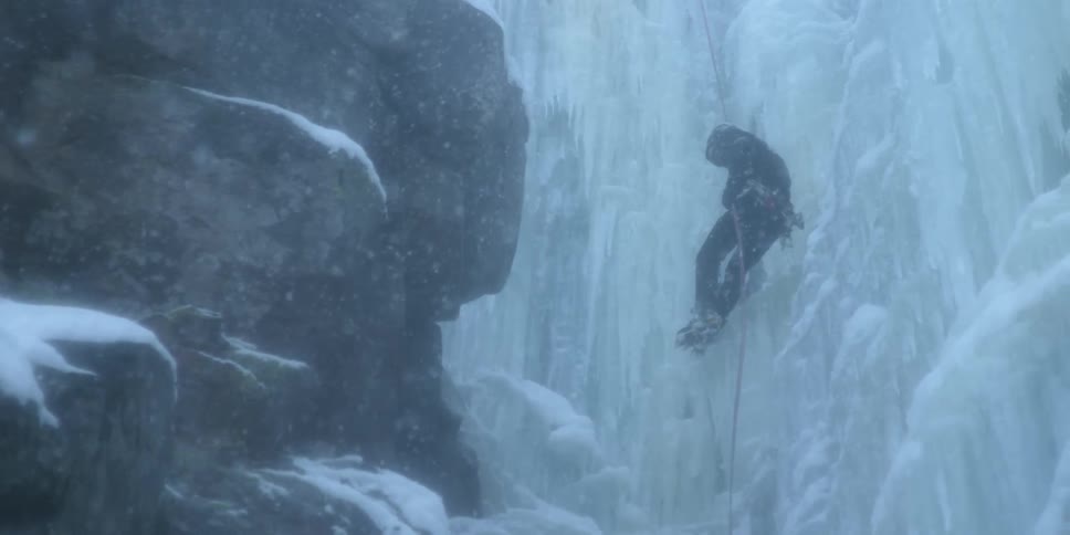 A man is climbing down a frozen waterfall as it snows