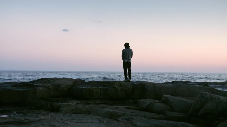 A person stands on some rocks by the shore as the sun sets