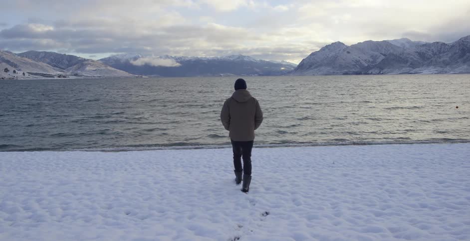 A man walks on snow towards a mountain lake
