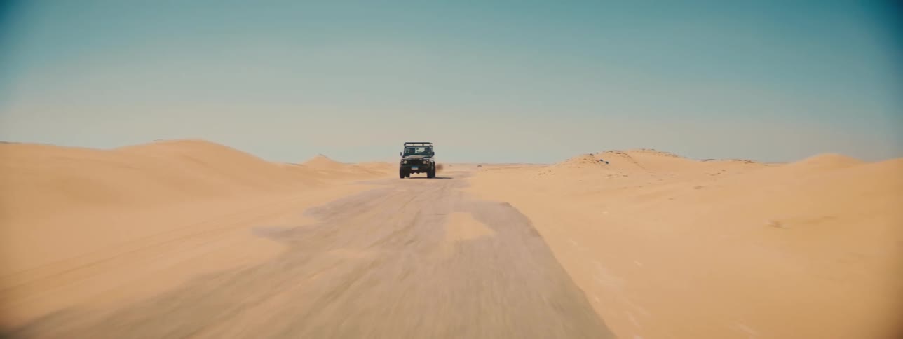A jeep drives down a deserted road past sand dunes in the desert