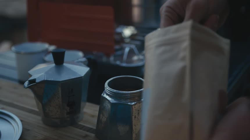 A woman is outside scooping coffee grinds from a pouch into a metal container with a camp stove in the background during the evening