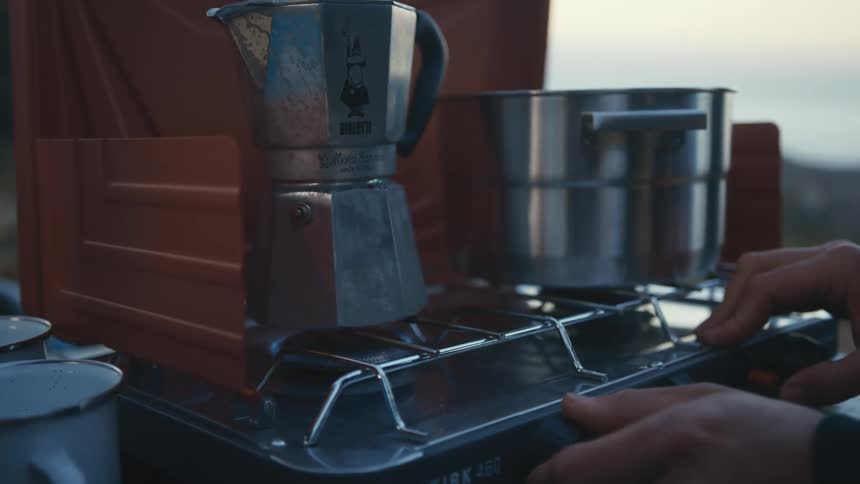 Someone turns on the flame under a teapot sitting next to a silver pot on a burner outside