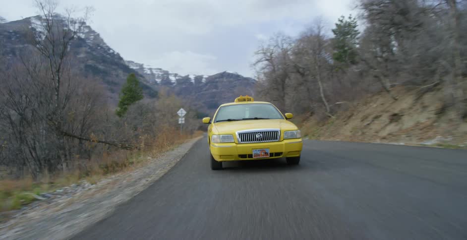 Taxi drives on country road next to trees with steep mountains in background on cloudy day